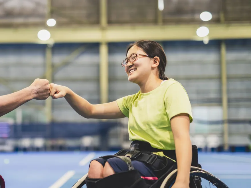 A young wheelchair user smiling and fist-bumping in a sports hall